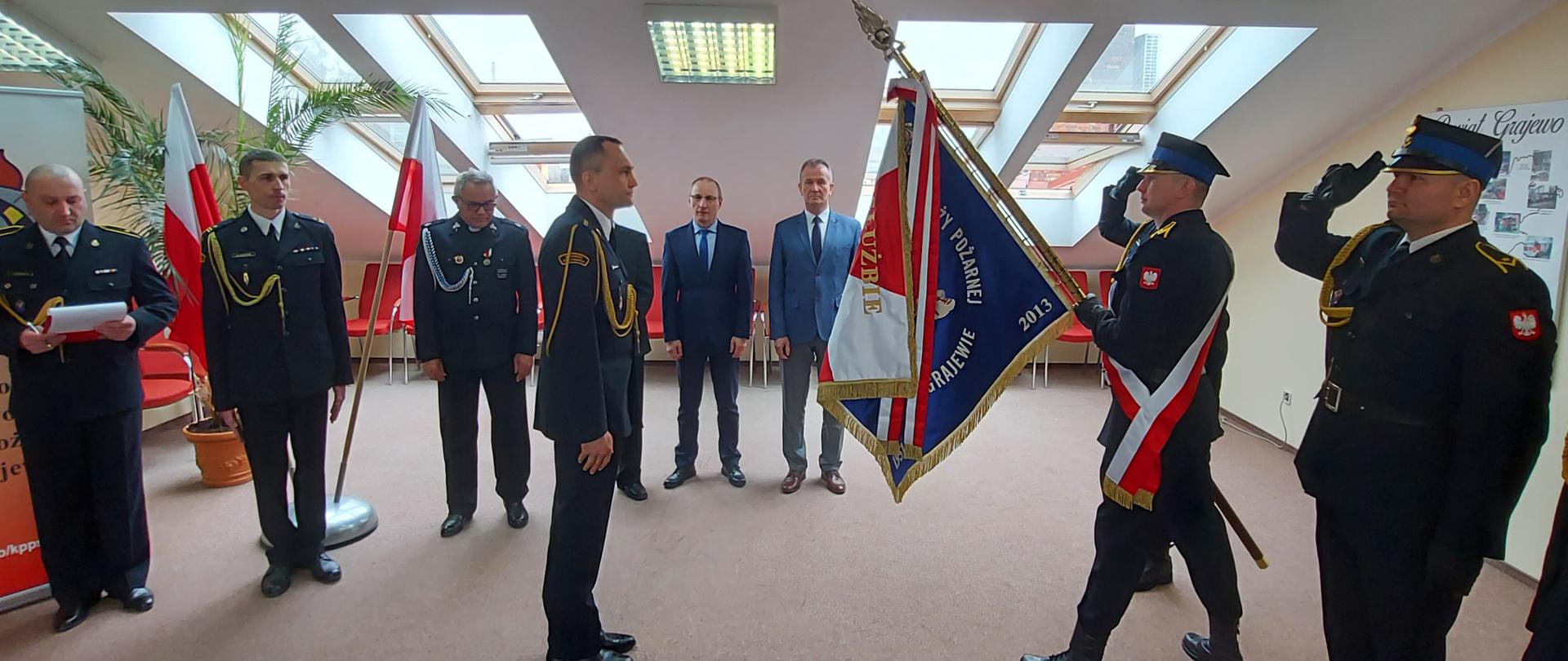 Nowy zastępca komendanta powiatowego PSP w Grajewie kpt. Jarosław Szeszko wita się ze sztandarem komendy. W tle stoją funkcjonariusze oraz zaproszeni goście. 
