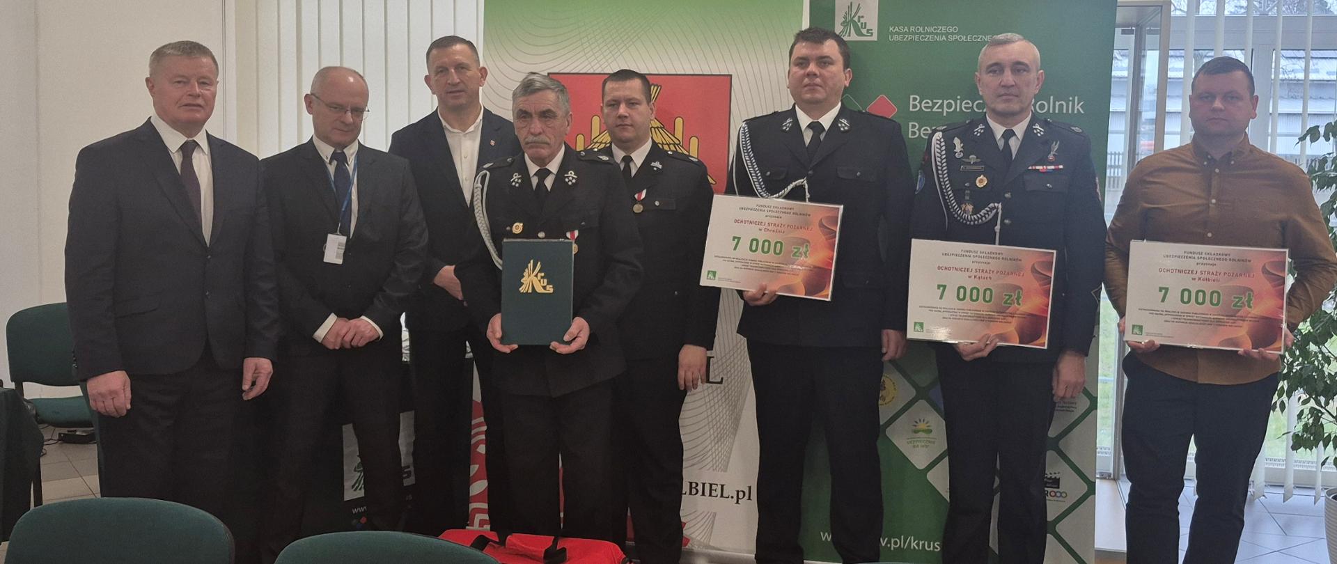 Ośmiu mężczyzn stoi tle banerów KRUS i Gminy Kołbiel, część z nich w galowych mundurach strażaka, trzymają plansze z kwotą otrzymanego wsparcia oraz potwierdzeniem otrzymania zestawu ratownictwa przedlekarskiego.
