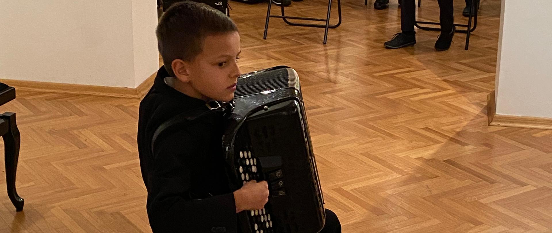 Zdjęcie przedstawiające akordeonistę z klasy mgr Mateusza Bassary. W tle inni akordeoniści słuchający występu kolegi podczas audycji klasy akordeonu. 