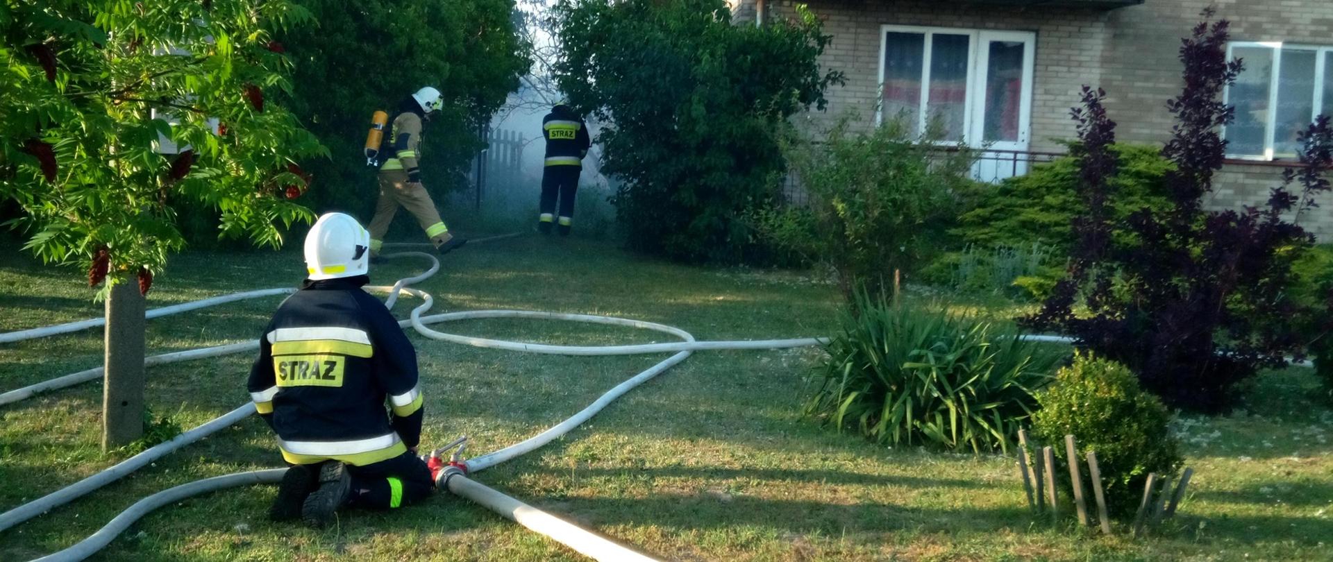 Zdjęcie przedstawia budynek mieszkalny, gdzie na poddaszu gospodarczym doszło do pożaru. Na trawniku przed domem rozwinięte linie wężowe, a przy rozdzielaczu klęczy ratownik. Dwóch ratowników widać w głębi podwórka. Po prawej stronie, u góry widać kosz drabiny mechanicznej z którego ratownicy podają prąd wody.