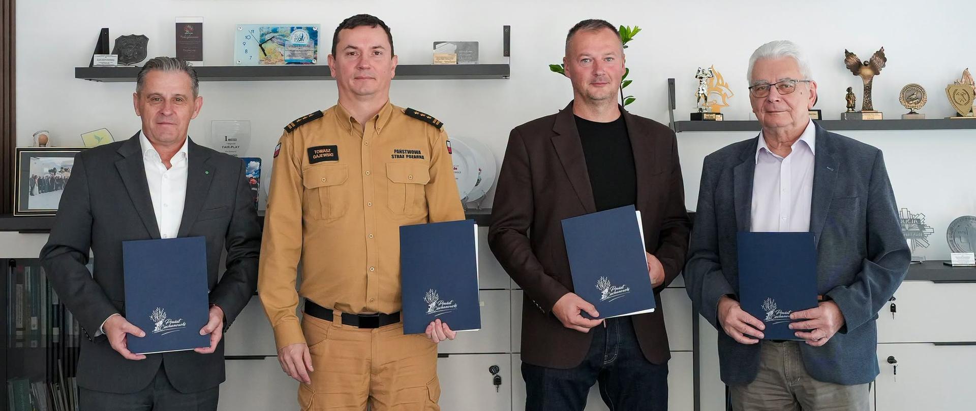 Cztery osoby stoją obok siebie i pozują do wspólnego zdjęcia. Komendant Powiatowy Państwowej Straży Pożarnej w Ciechanowie oraz trzej mężczyźni w ubraniach cywilnych trzymają granatowe teczki z dokumentami. W tle znajdują się półki z pamiątkami, dyplomami i statuetkami.