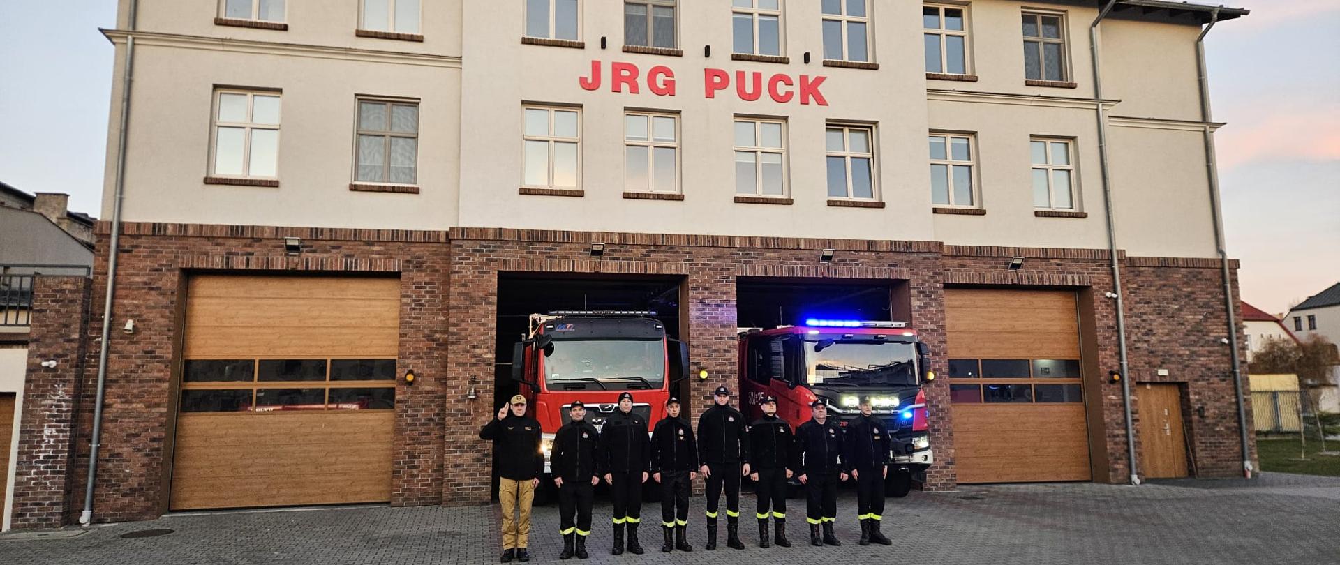 Strażacy ustawieni w szeregu oddają honor zmarłemu koledze. Na drugim planie budynek Jednostki Ratowniczo Gaśniczej oraz wozy bojowe z włączonymi sygnałami świetlno - dźwiękowymi.