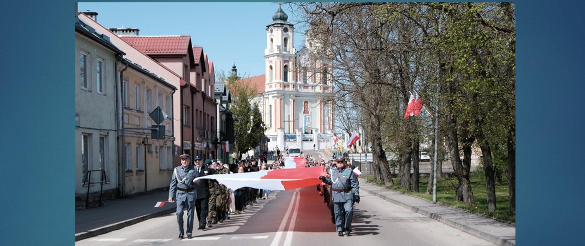 Uczestnicy uroczystości podczas przemarszu przez miasto niosą biało-czerwoną flagę.