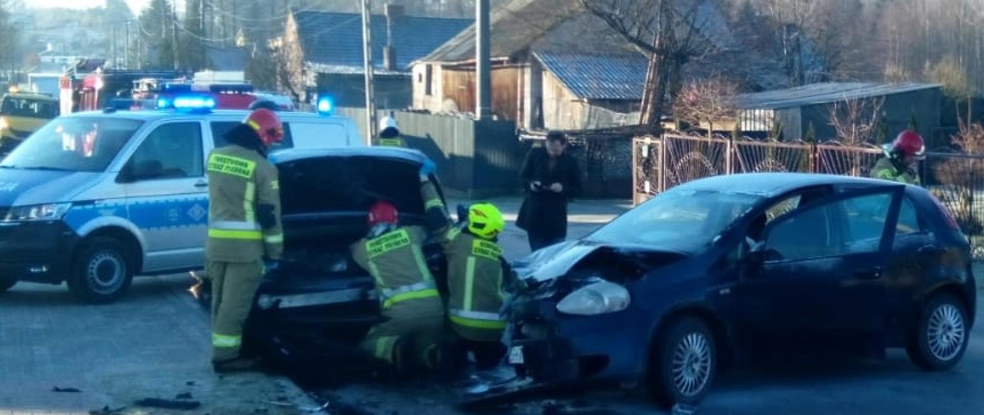 Zdjęcie przedstawia dwa samochody osobowe, które uległy zderzeniu, przy których pracuje trzech strażaków. W tle wiać radiowóz policyjny.