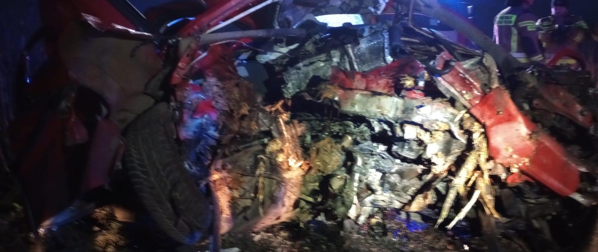 Zdjęcie przedstawia samochód, z rozbitym przodem. Obok stoją strażacy.