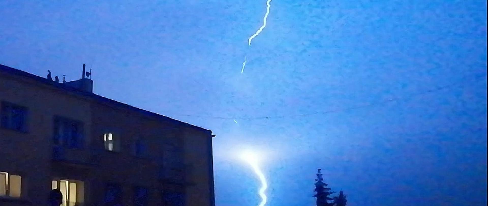 Zdjęcie przedstawia wyładowanie atmosferyczne w tle budynek wielorodzinny
