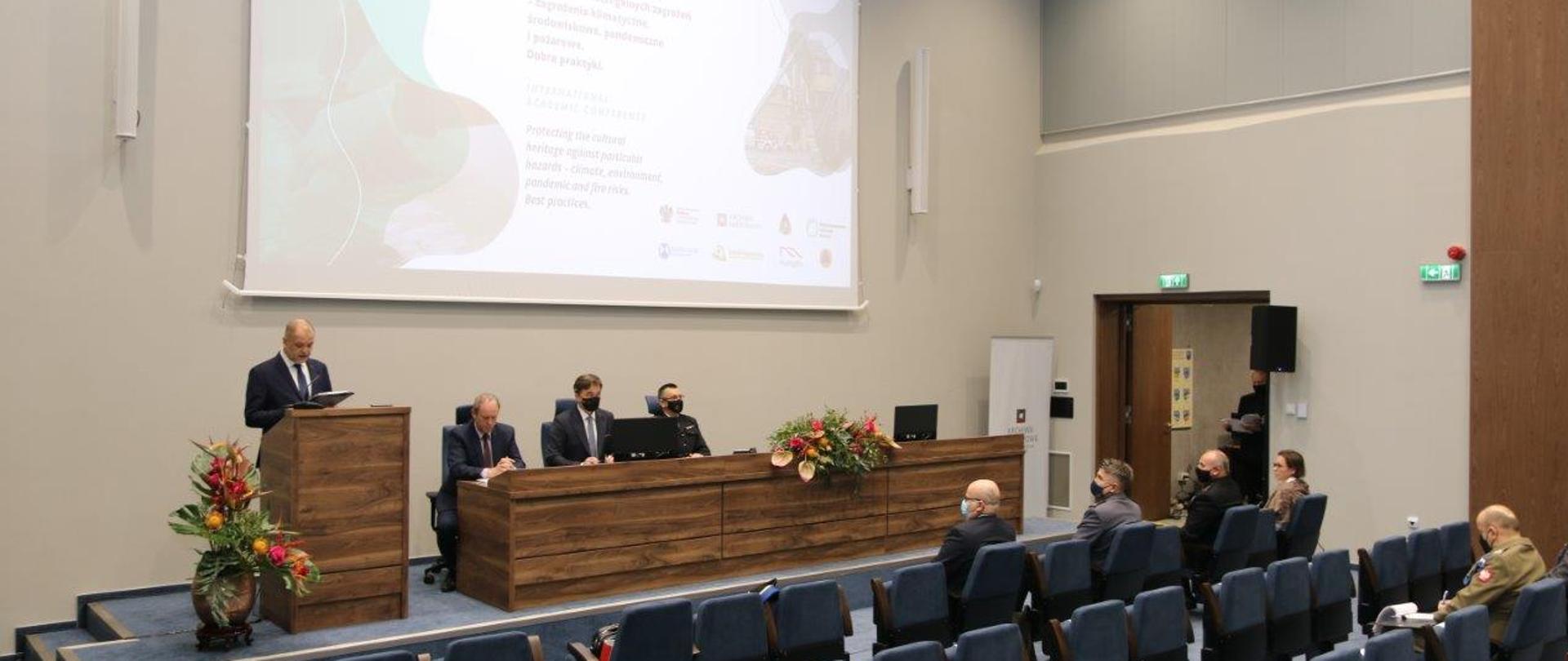 Widok na salę w trakcie konferencji. Prelegent stojący przy mównicy, reszta uczestników rozsadzona zgodnie z zasadami reżimu sanitarnego.