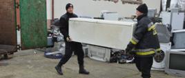 Strażacy dla Tosi. Plac komendy w Rawiczu. Na nim złożone są elektrośmieci w postaci m.in lodówek, monitorów komputerowych, drukarek, itp. Dwóch strażaków przenosi lodówkę do podstawionego kontenera.