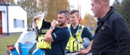 dwaj policjanci prezentują dzieciom kamizelki policyjne i co się znajduje w poszczególnych kieszonkach za nimi znajduje się niebiesko żółty motocykl