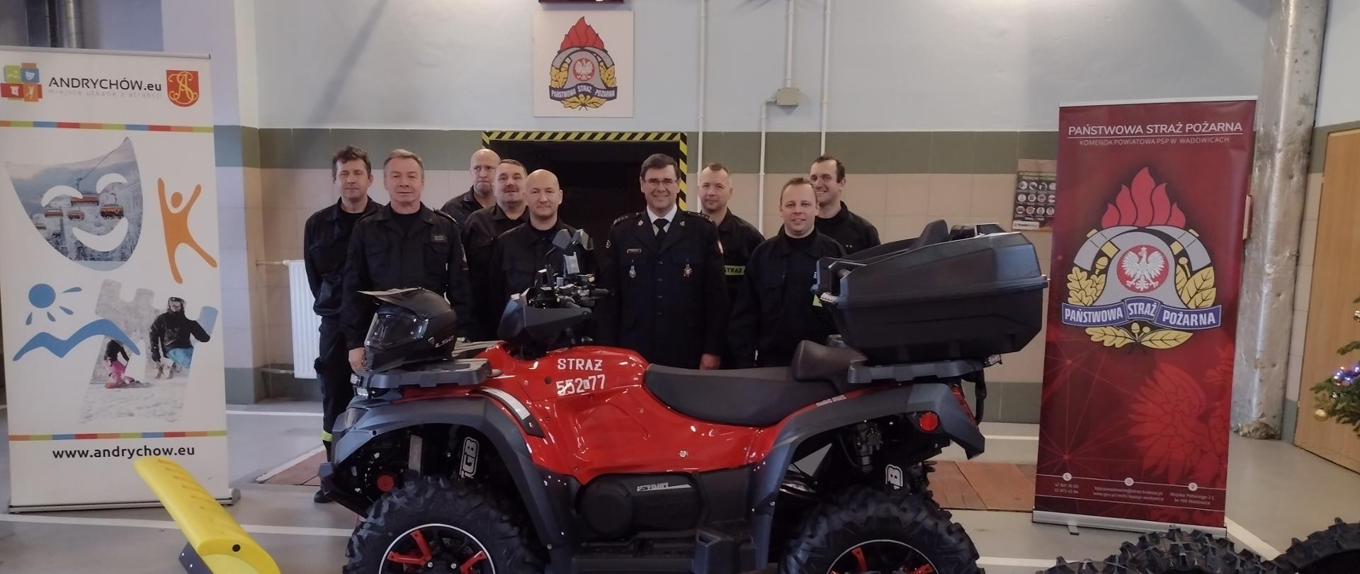 Na pierwszym planie quad z dodatkowym wyposażeniem. Za quadem Komendant w mundurze wyjściowym oraz 8 strażaków w mundurach koszarowych.Na bokach rollupy. Po prawej stronie rollup PSP, po lewej Gminy Andrychów