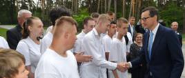 Na dworze, premier Morawiecki podaje rękę młodemu człowiekowi w białej koszuli, obok stoi kilkanaście osób, w tle zielone drzewa.