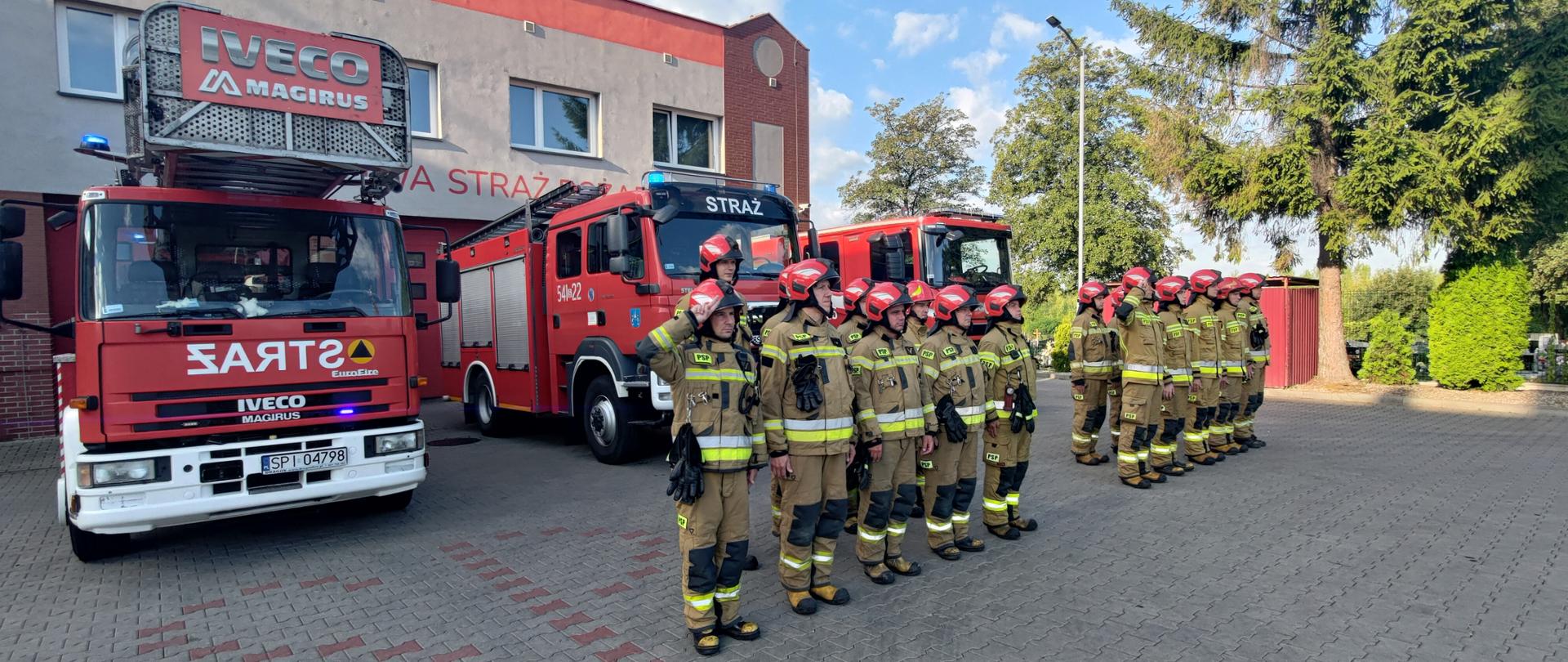 Strażacy w ubraniach specjalnych stoją w dwóch pododdziałach na tle pojazdów pożarniczych