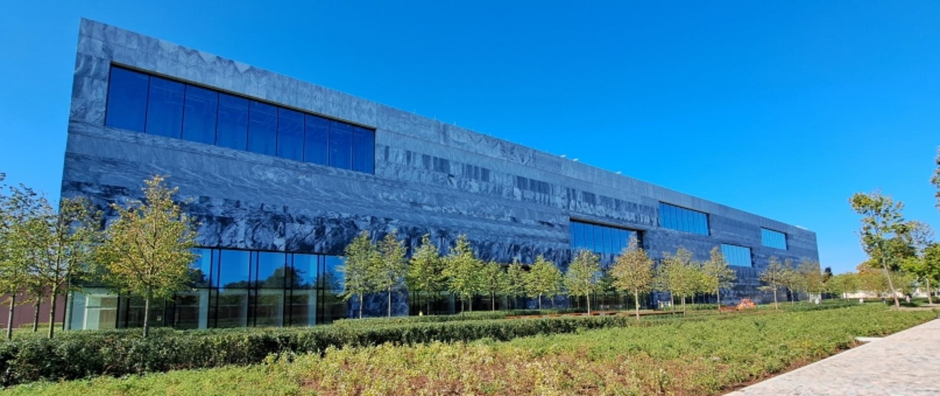 Nowoczesny budynek z dużymi oknami, otoczony zielenią, w słoneczny dzień.