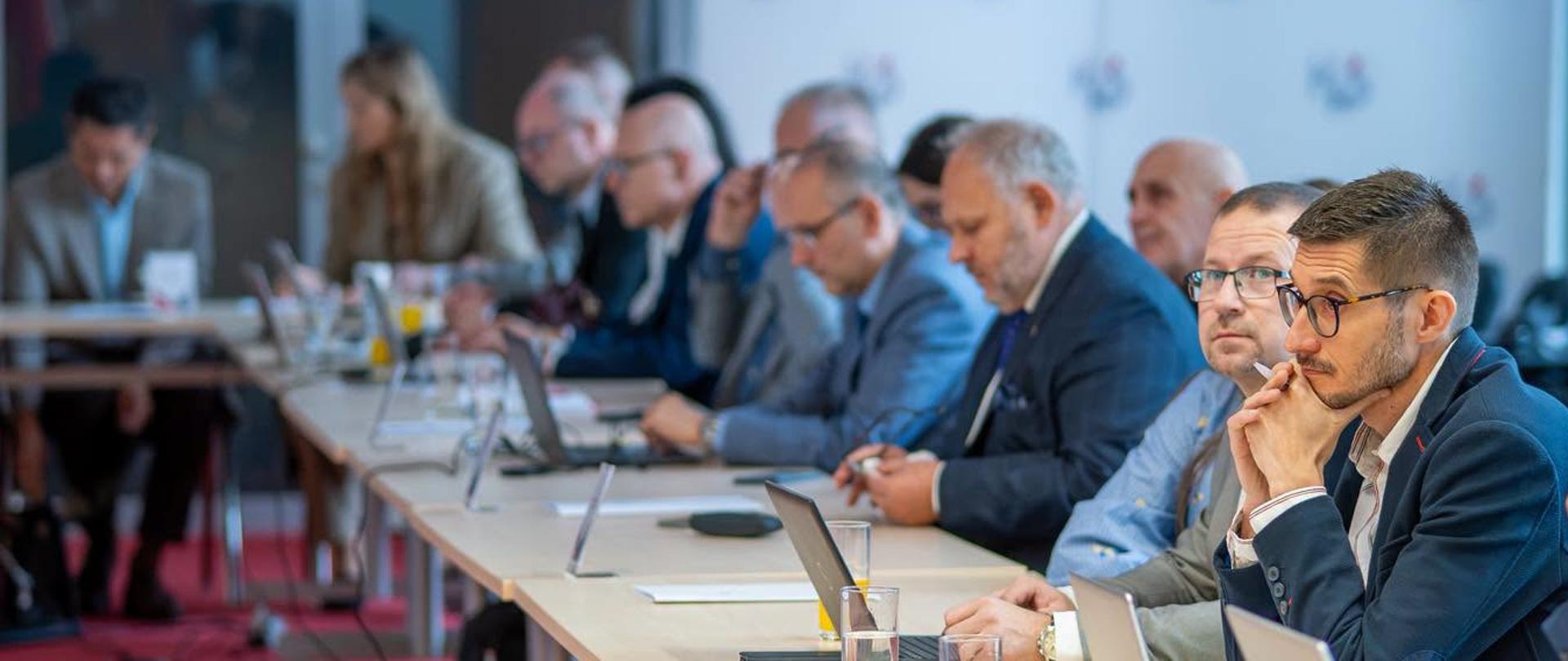 Kilkanaście osób siedzi przy ustawionych w podkowę stołach konferencyjnych. Na stołach znajdują się laptopy, dokumenty oraz szklanki z napojami.