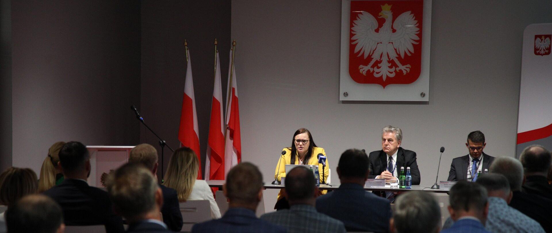 Na sali siedzi tyłem grupa ludzi. Przodem do nich przy stole siedzi od lewej minister M. Jarosińska-Jedynak