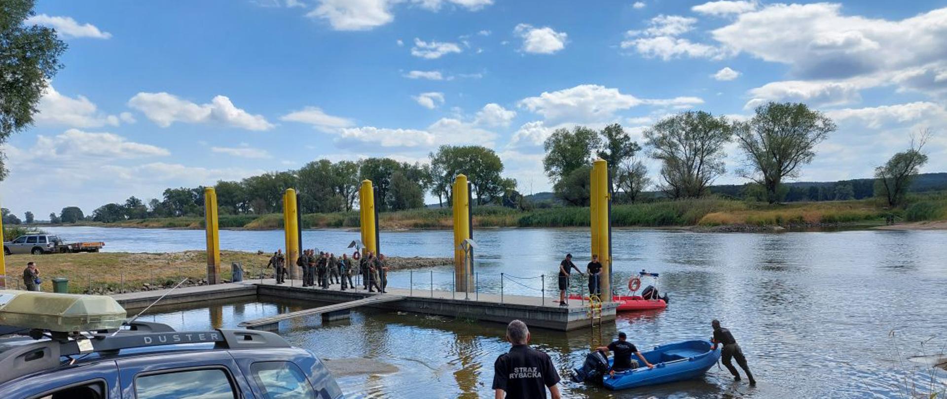 Zdjęcie przedstawia przygotowanie do podjęcia działań mających na celu patrolowanie oraz oczyszczenie rzeki Odry. Na zdjęciu widać funkcjonariuszy Państwowej Straży Pożarnej, żołnierzy Wojsko Polskie oraz przedstawicieli Straży Rybackej na tle portu w miejscowości Gostchorze.