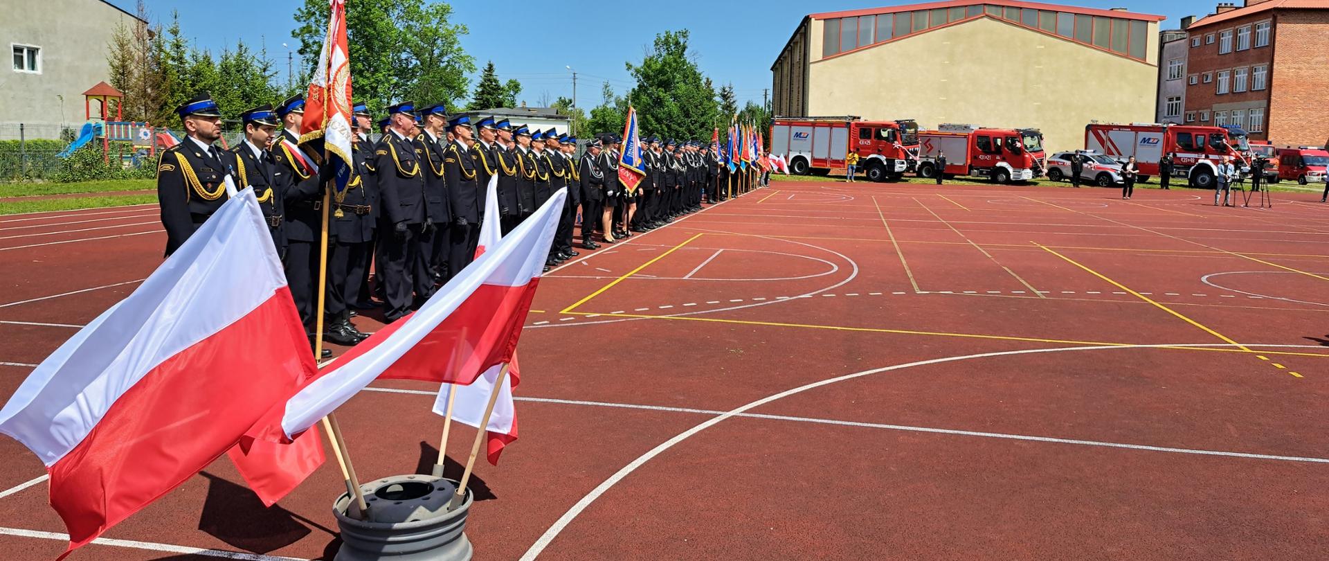 Zdjęcie przedstawia boisko szkolne przy szkole w Widawie. Na pierwszym planie powiewają polskie flagi, dalej stoją strażacy, samochody oraz zaproszeni goście. 