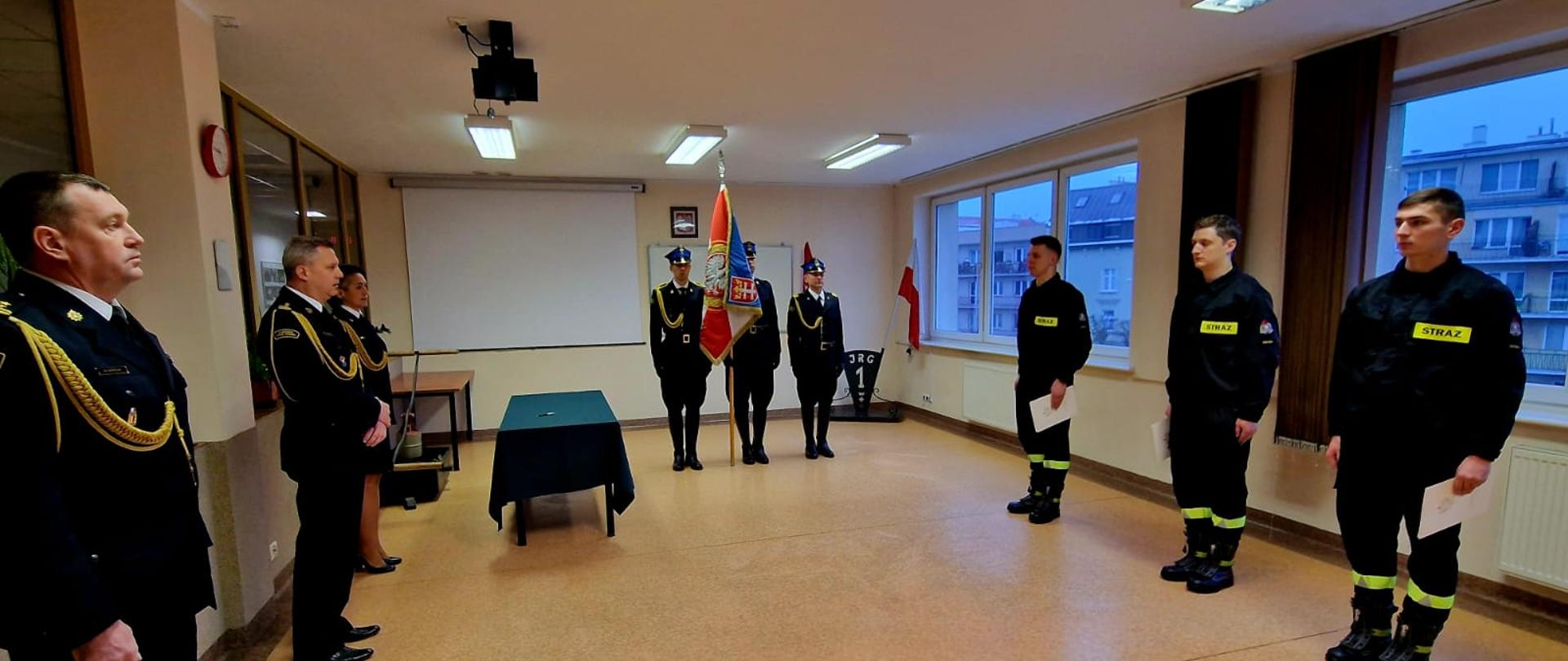 Na świetlicy JRG 1 podczas uroczystej zbiórki stoją: komendant miejski PSP, dowódca JRG, poczet sztandarowy oraz 3 funkcjonariuszy w ubraniu koszarowym.