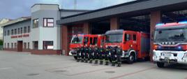 Grupa strażaków w czarnych ubraniach koszarowych z zielonymi odblaskami stoli w szeregu na tle samochodów pożarniczych. Na drugim planie budynek straży pożarnej. 