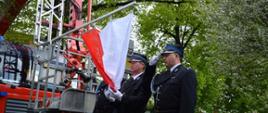 Narodowe Święto Trzeciego Maja. Plac Wolności w Rawiczu. Poczet flagowy z OSP dokonuje podniesienia flagi państwowej. Flaga jest przymocowana do kosza specjalnego wozu strażackiego z podnośnikiem hydraulicznym. Strażak stojący z prawej strony salutuje, natomiast strażak stojący w środku - przytrzymuje flagę. W tle drzewa i budynki.