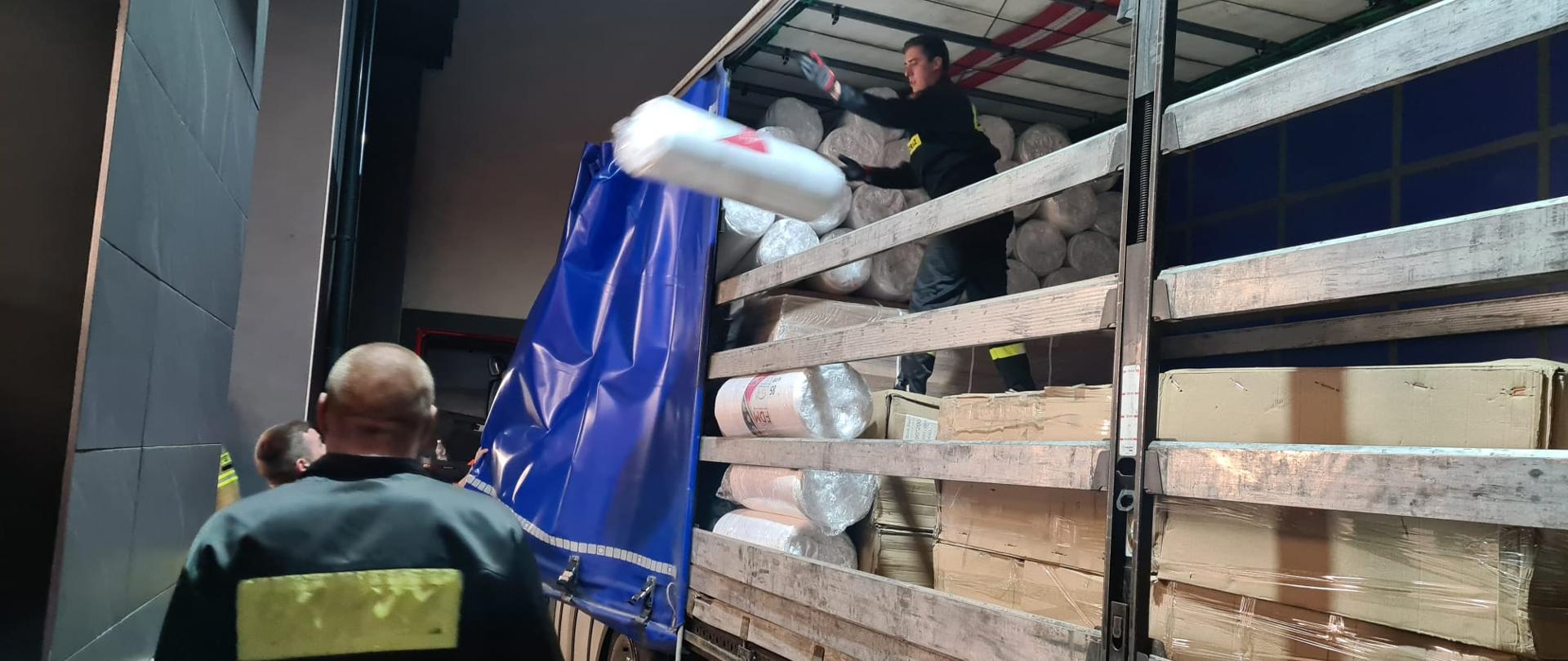 Strażacy rozładowują przywiezione materiały z samochodu ciężarowego. 