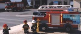 na pierwszym planie znajduje się czerwony samochód strażacki z białą drabiną na dachu stojący na jasnym kamieniu wykonany z klocków lego, obok samochodu stoją trzy ludziki z klocków lego strażacy. w tle zdjęcia znajduje się remiza ochotniczej straży pożarnej obok której stoi czerwony samochód pożarniczy