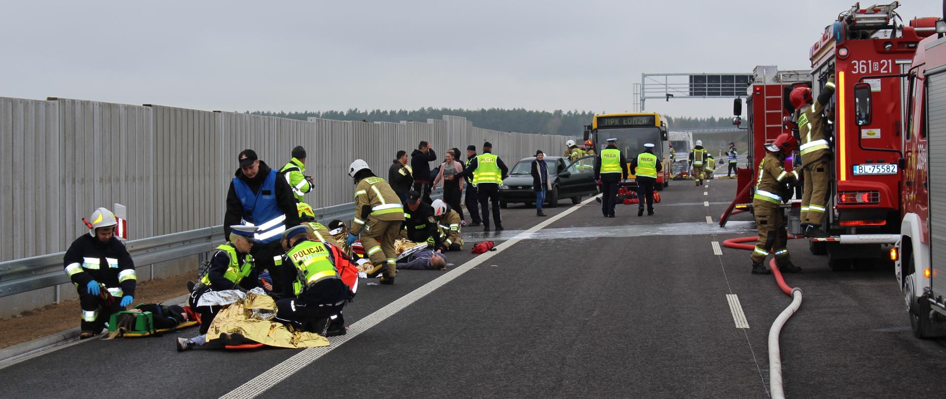 Strażacy i policjanci udzielają pomocy poszkodowanym leżącym na drodze. W tle samochody strażackie.