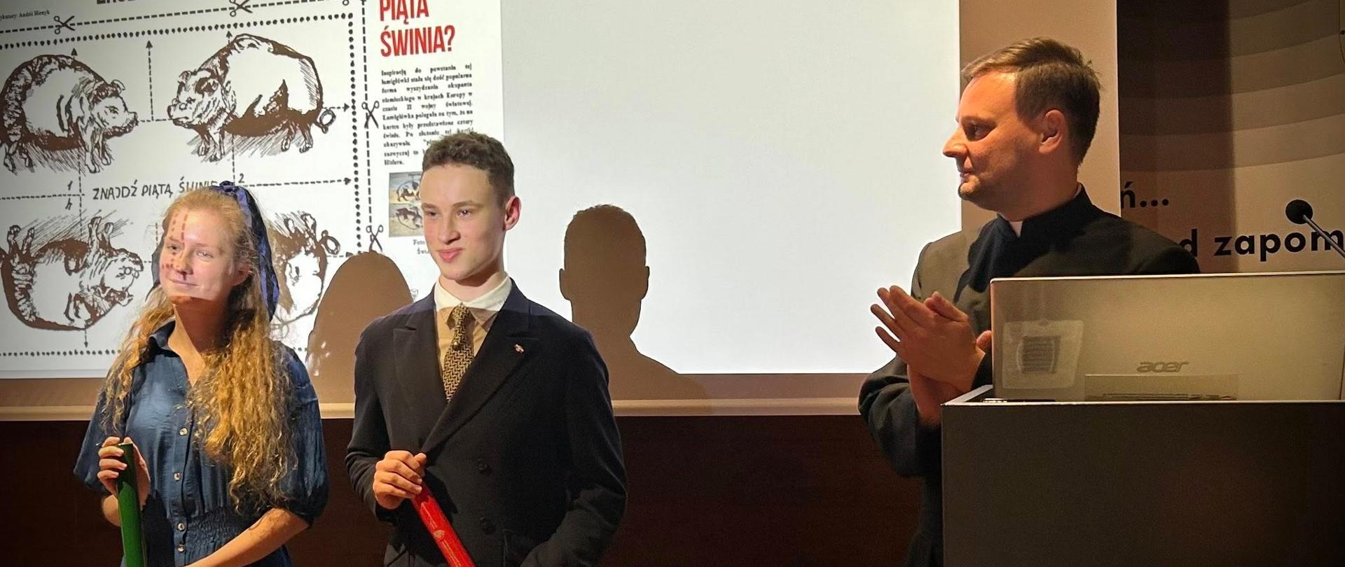 Na zdjęciu widać trzy osoby stojące w sali podczas uroczystości w Muzeum Katyńskim. Po lewej stronie znajdują się dwie młode osoby, Paulina Nadworska, Andrii Hirnyk trzymające w dłoniach zwinięte dyplomy – kobieta w niebieskiej sukience i mężczyzna w garniturze. Po prawej stronie stoi mężczyzna w ciemnym stroju, bijący brawo. W tle widoczny jest fragment prezentacji z napisem „Kim jest piąta świnia?” oraz logo MK Muzeum Katyńskie na mównicy. Scena przedstawia moment wręczenia nagród lub wyróżnień w konkursie historycznym.