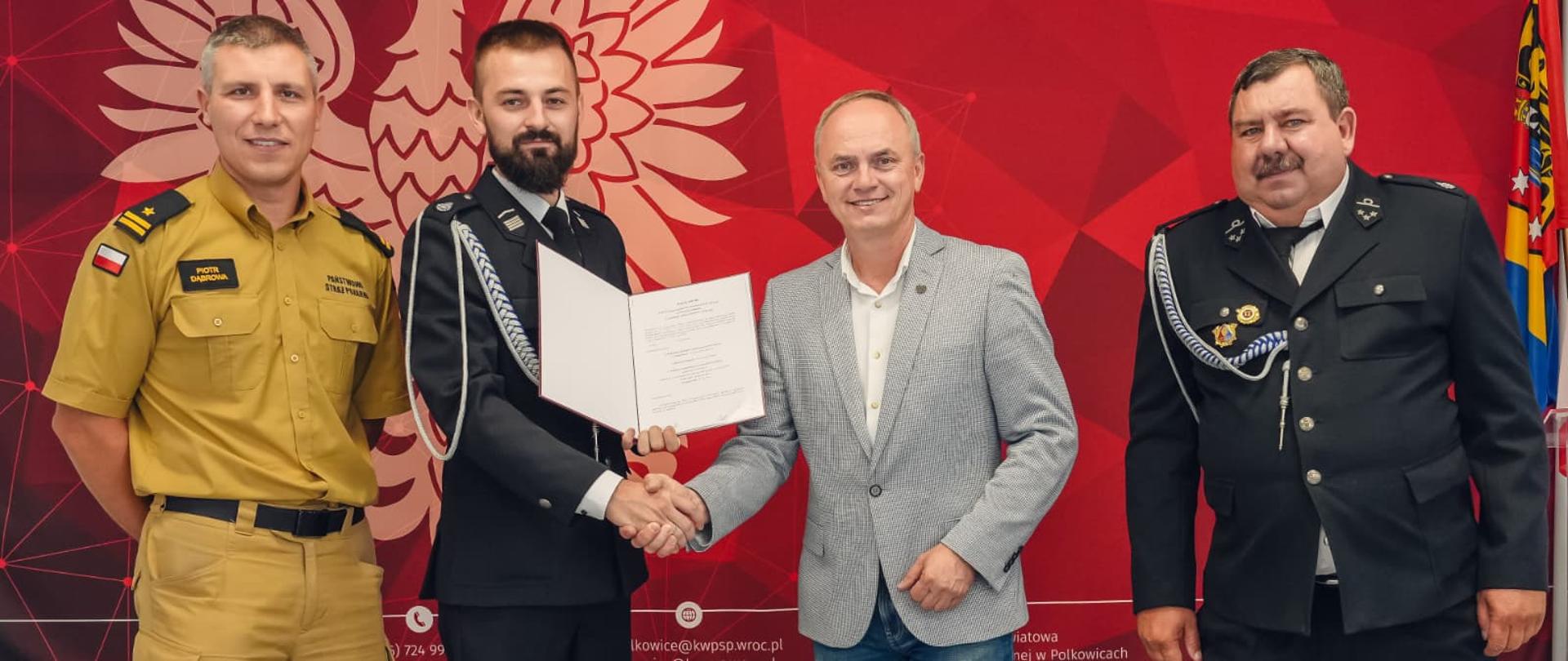 Zdjęcie przedstawia oficjalne spotkanie lub uroczystość wręczenia dokumentu/aktu nominacji w jednostce Straży Pożarnej.
Główne elementy zdjęcia
• Uczestnicy: * Na zdjęciu znajduje się czterech mężczyzn stojących obok siebie.
o Trzech z nich ma na sobie mundury (zróżnicowane pod względem typu i stopnia służbowego) związane z Państwową Strażą Pożarną (PSP) lub strażą ochotniczą.
o Jeden z mężczyzn (drugi od prawej) ubrany jest w czarno-białą stylizację cywilną/półoficjalną: szarą marynarkę, białą koszulę i dżinsy.
• Moment przekazania:
o Mężczyzna w ciemnym mundurze galowym (drugi od lewej) potrząsa dłonią mężczyzny w szarej marynarce.
o Jednocześnie trzyma otwartą czerwoną teczkę z oficjalnym pismem/dokumentem skierowanym do wglądu.
Tło i otoczenie
• Ścianka reklamowa/tło: W tle znajduje się duża, czerwona ścianka z godłem Polski (Orłem Białym), logo Państwowej Straży Pożarnej oraz napisem:
PAŃSTWOWA STRAŻ POŻARNA > KOMENDA POWIATOWA PSP W POLKOWICACH
• Flaga: Po prawej stronie widoczny jest fragment podwieszonej flagi lub proporca.
