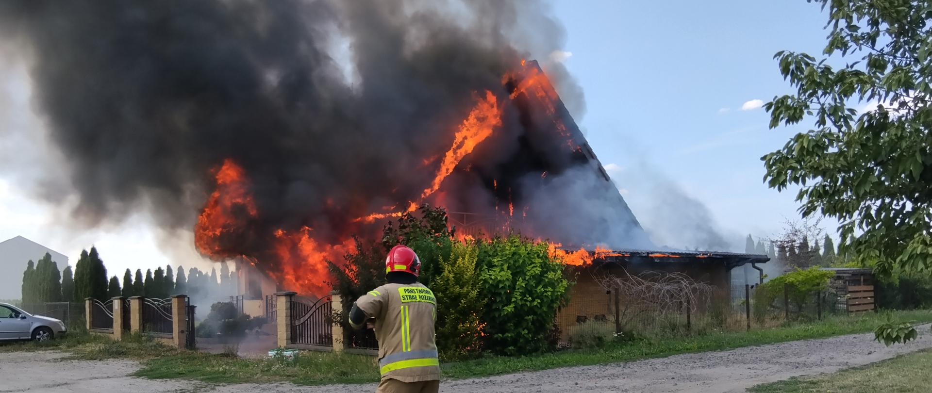 Pożar budynku mieszkalnego jednorodzinnego. Nad budynkiem unosi się gęsty dym. i płomienie. Na zdjęciu widać strażaka. 