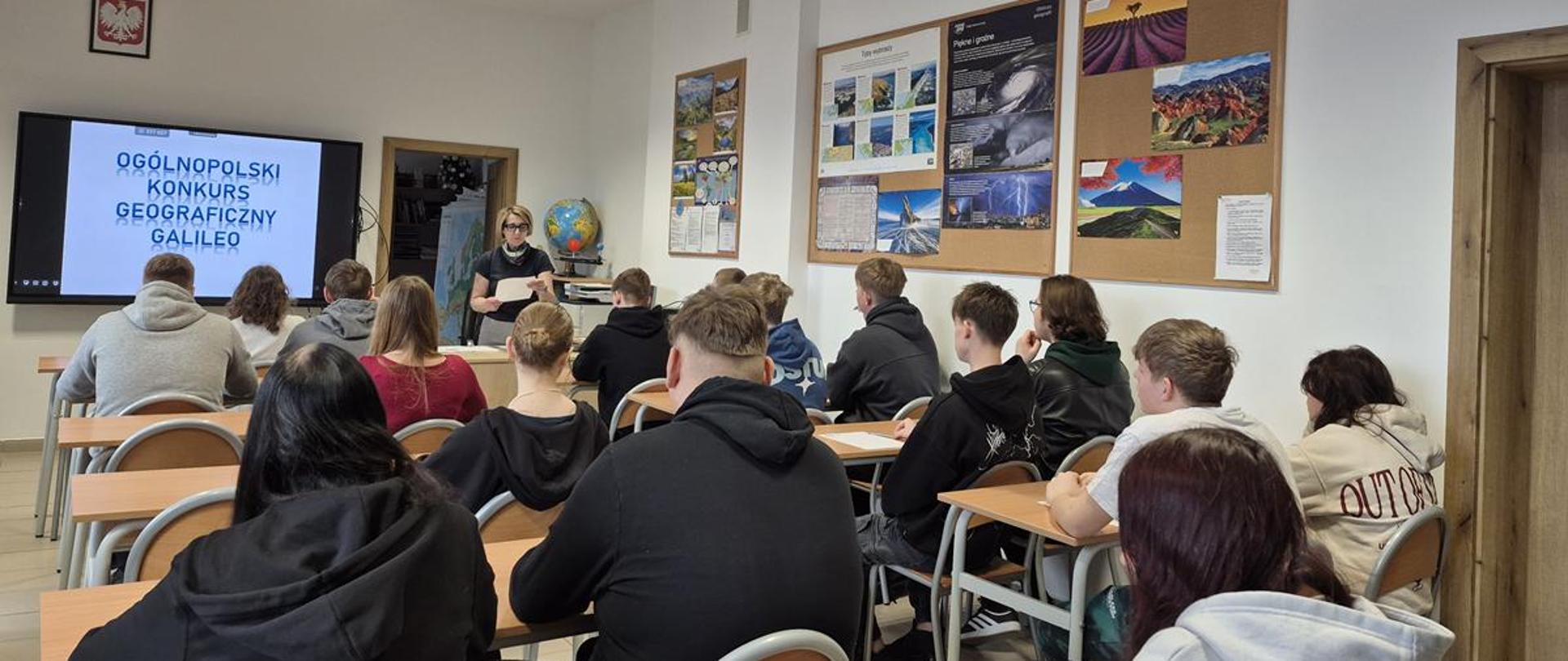 Uczniowie siedzą w klasie, przed nimi stoi nauczycielka, na monitorze wyświetlony jest napis "Ogólnopolski Konkurs Geograficzny GALILEO".