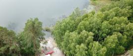 Zdjęcie wykonane z powietrza za pomocą drona, przedstawiające punkt czerpania wody, przenośny zbiornik na wodę, samochód pożarniczy należący do KP PSP Chojnice oraz linię gaśniczą rozwiniętą wzdłuż leśnej drogi.