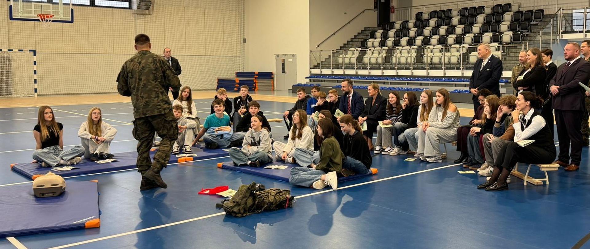 sala sportowa, w niej żołnierz uczący dzieci pierwszej pomocy, na ławkach obok siedzą ministrowie i wojewoda