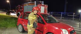 Strażak stoi obok uszkodzonego samochodu osobowego koloru czerwonego. W tle samochód pożarniczy.