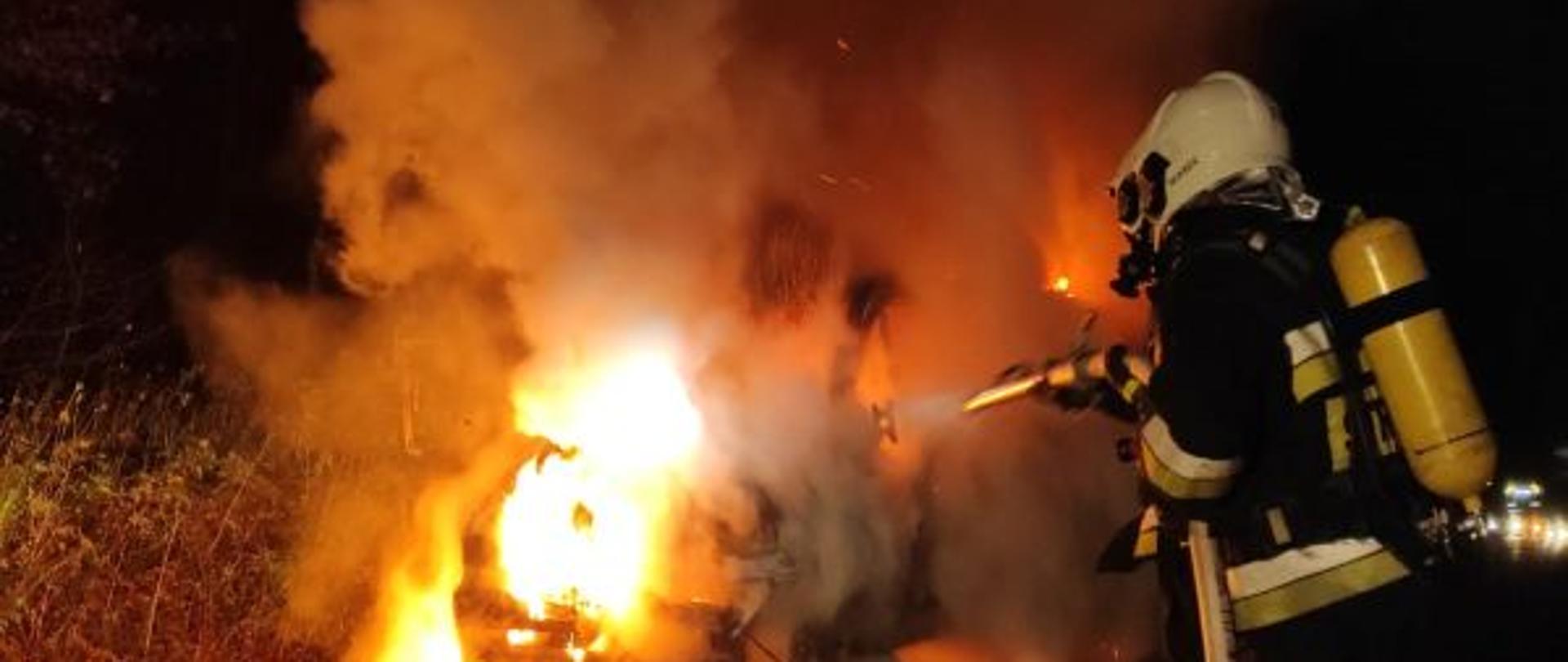 Zdjęcie wykonane w nocy. Oświetlone przez palący się pojazd. Samochód dostawczy całkowicie objęty ogniem. Przy aucie strażacki w ubraniu bojowym , w sprzęcie ochrony dróg oddechowych. Strażak trzyma prądownicę i wąż. Gasi palący się pojazd