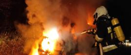 Zdjęcie wykonane w nocy. Oświetlone przez palący się pojazd. Samochód dostawczy całkowicie objęty ogniem. Przy aucie strażacki w ubraniu bojowym , w sprzęcie ochrony dróg oddechowych. Strażak trzyma prądownicę i wąż. Gasi palący się pojazd