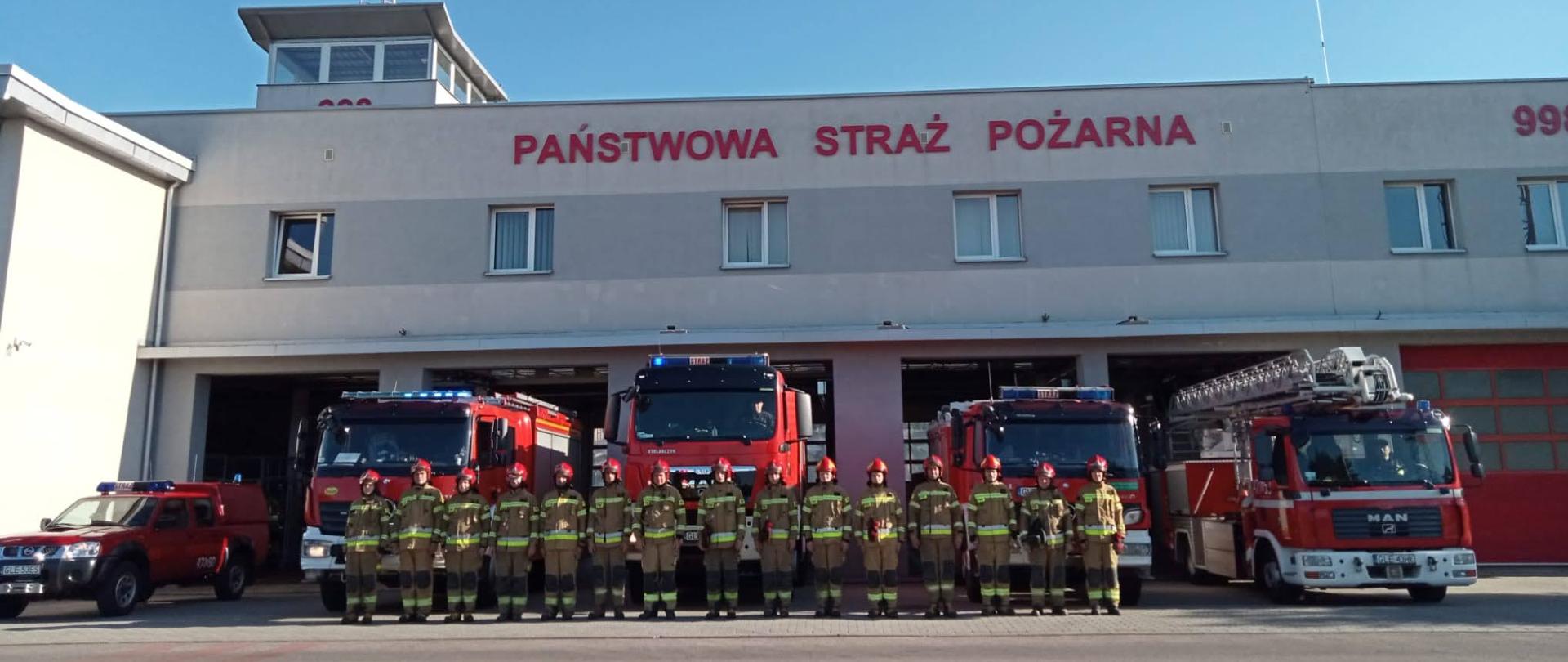 Strażacy Państwowej Straży Pożarnej stoją przed samochodami gaśniczymi oddając hołd tragicznie zmarłemu strażakowi z Komendy Głównej Państwowej Straży Pożarnej. 