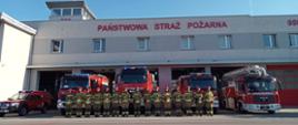 Strażacy Państwowej Straży Pożarnej stoją przed samochodami gaśniczymi oddając hołd tragicznie zmarłemu strażakowi z Komendy Głównej Państwowej Straży Pożarnej. 