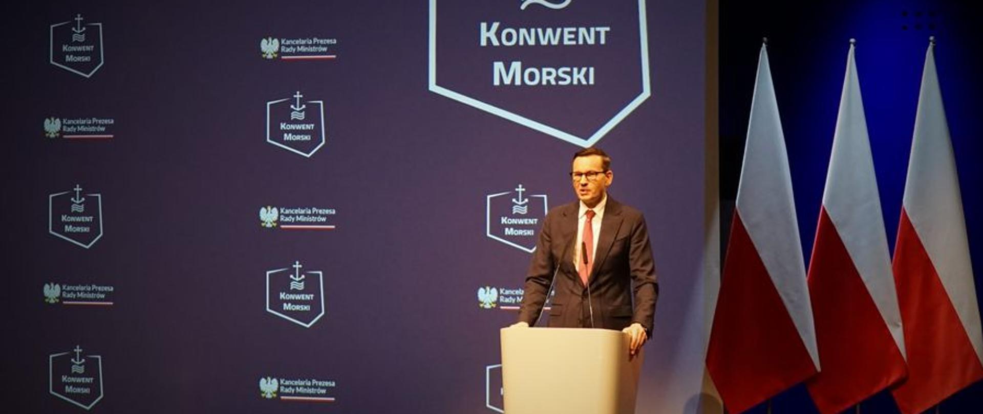Mężczyzna stojąc przy mównicy przemawia za nim ustawione są trzy flagi Polski oraz baner z napisem Konwent Morski oraz logo.