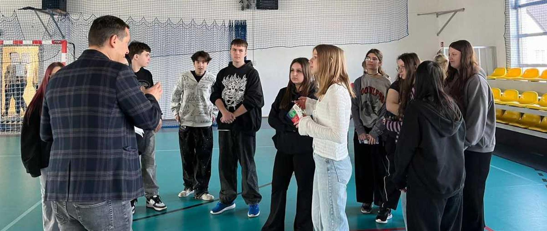 Ta fotografia przedstawia grupę młodych ludzi wewnątrz sali gimnastycznej.
Grupa uczniów zgromadziła się wokół osoby dorosłej w celu rozmowy lub instruktażu.
W tle widoczne są trybuny z żółtymi siedzeniami oraz siatki zabezpieczające.
Obiekt znajduje się we Włocławku i jest częścią lokalnej infrastruktury edukacyjnej.
Tego typu spotkania w szkołach często służą aktywnościom edukacyjnym lub sportowym.
