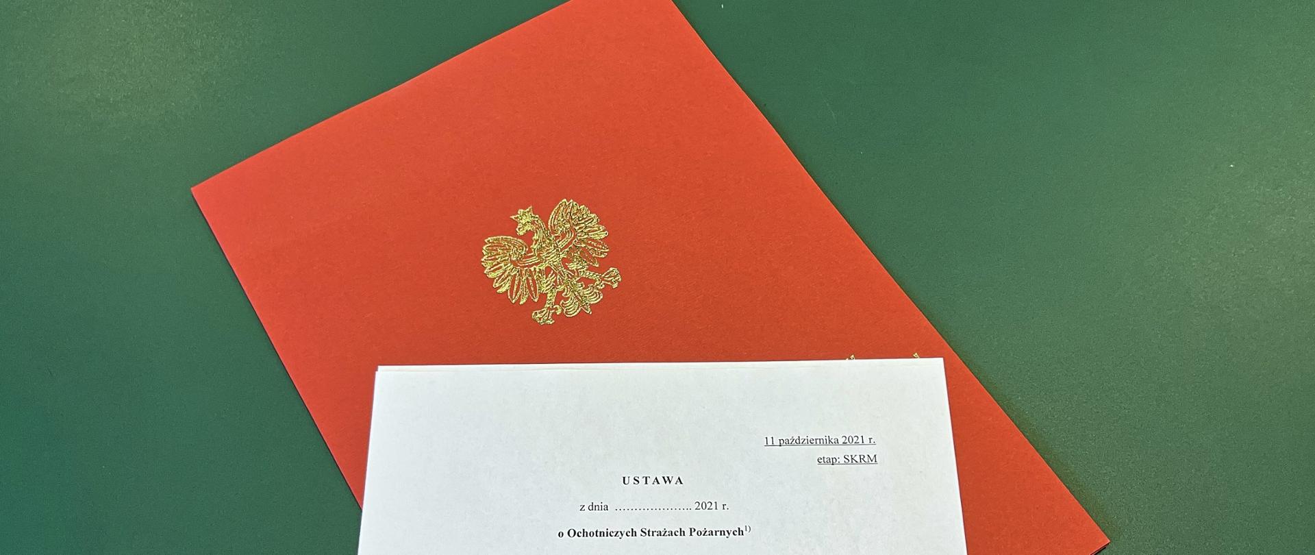 Biała kartka z tytułem dokumentu ułożona na czerwonej teczce papierowej z orłem, pod spodem zielone tło