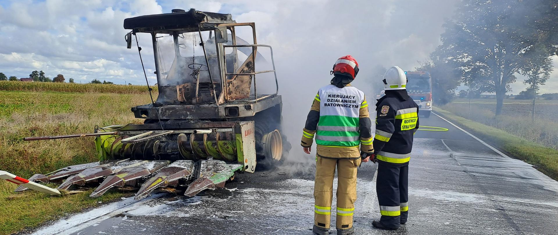 Zdjęcie drogę krajową na której stoi palący się kombajn do koszenia kukurydzy oraz strażaków podczas prowadzenia akcji gaśniczej.