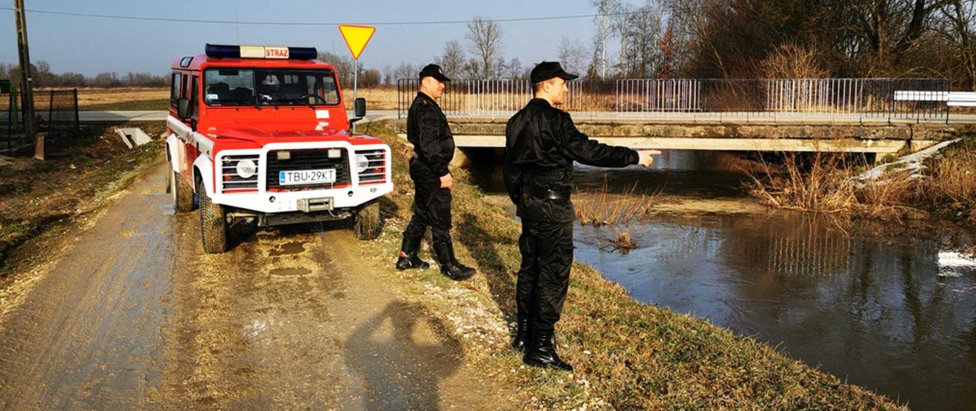 Na zdjęciu dwóch strażaków stojących na brzegu rzeki i monitorujących ciek wodny, w tle samochód strażacki terenowy w kolorze czerwonym.