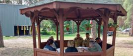 Wizytacja obozu ZHP w Białym Brzegu, pod drewnianą altaną siedzą przedstawiciele służb odpowiedzialni z bezpieczeństwo w trakcie trwania letniego wypoczynku