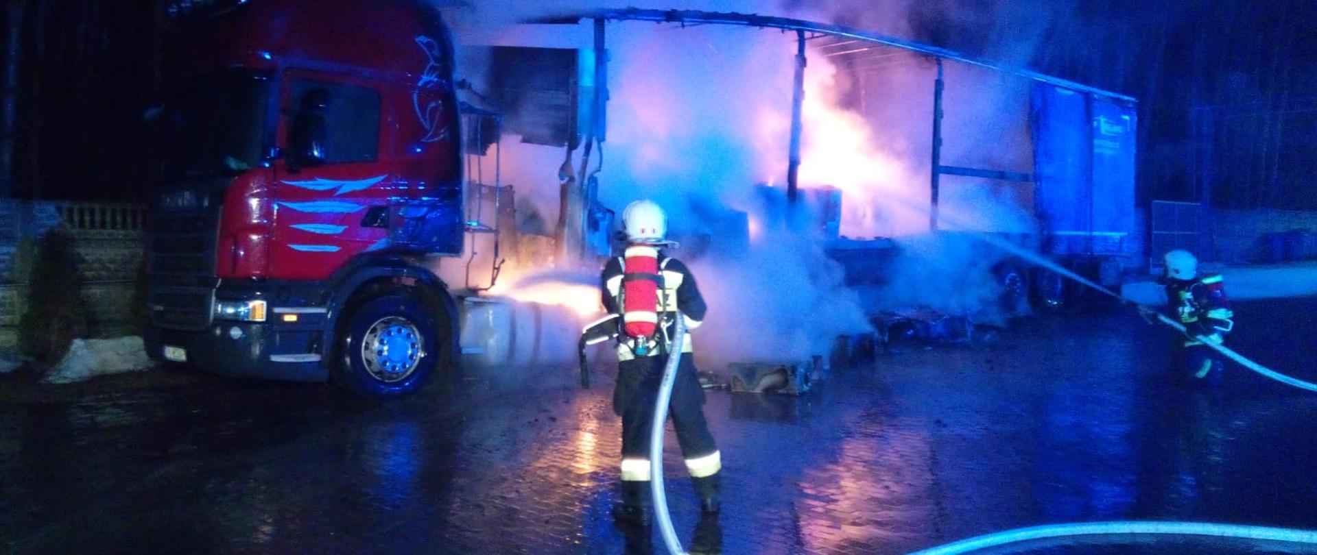 Fotografia przedstawia dwóch strażaków gaszących palaczy się samochód ciężarowy i naczepę