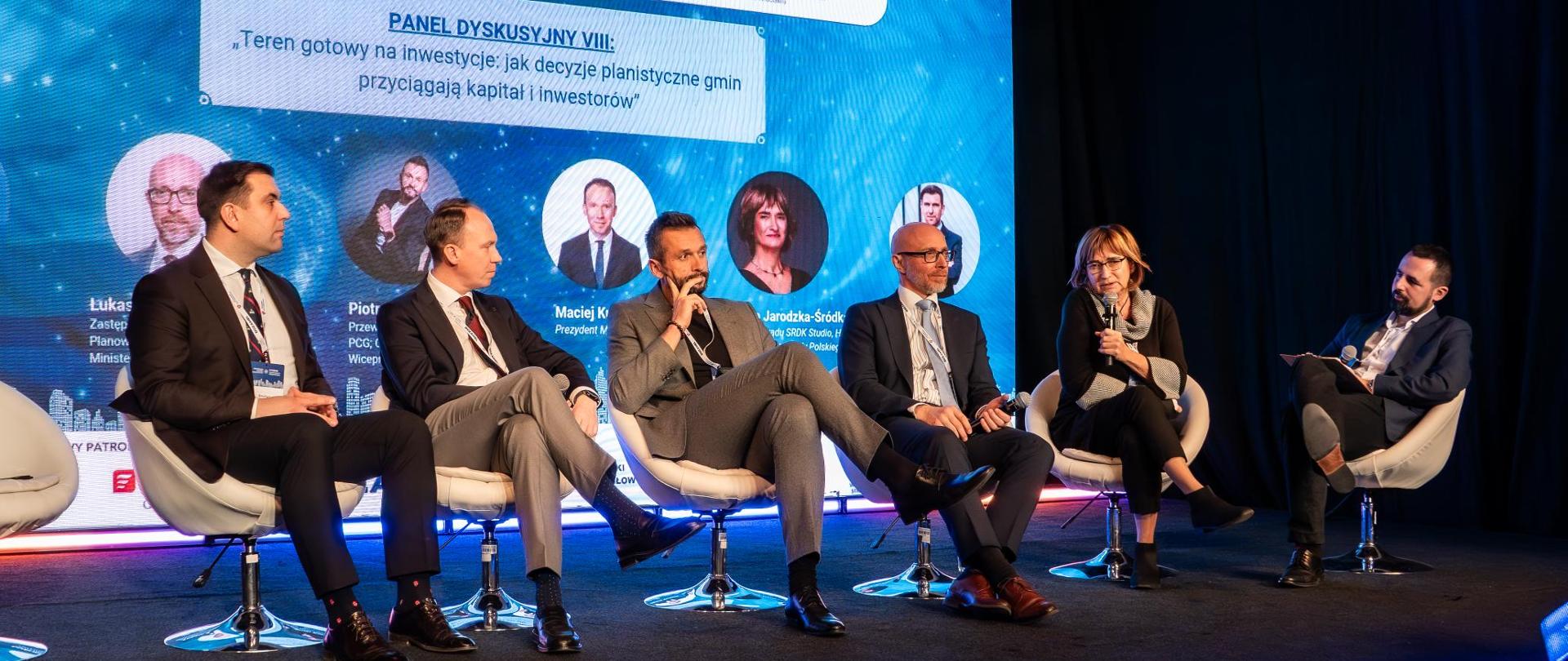 Zdjęcie przestawia scenę, na której zasiada sześciu prelegentów. Za prelegentami na ekranie ledowym wyświetlony jest tytuł panelu dyskusyjnego "Teren gotowy na inwestycje: jak decyzje planistyczne gmin przyciągają kapitał i inwestorów".