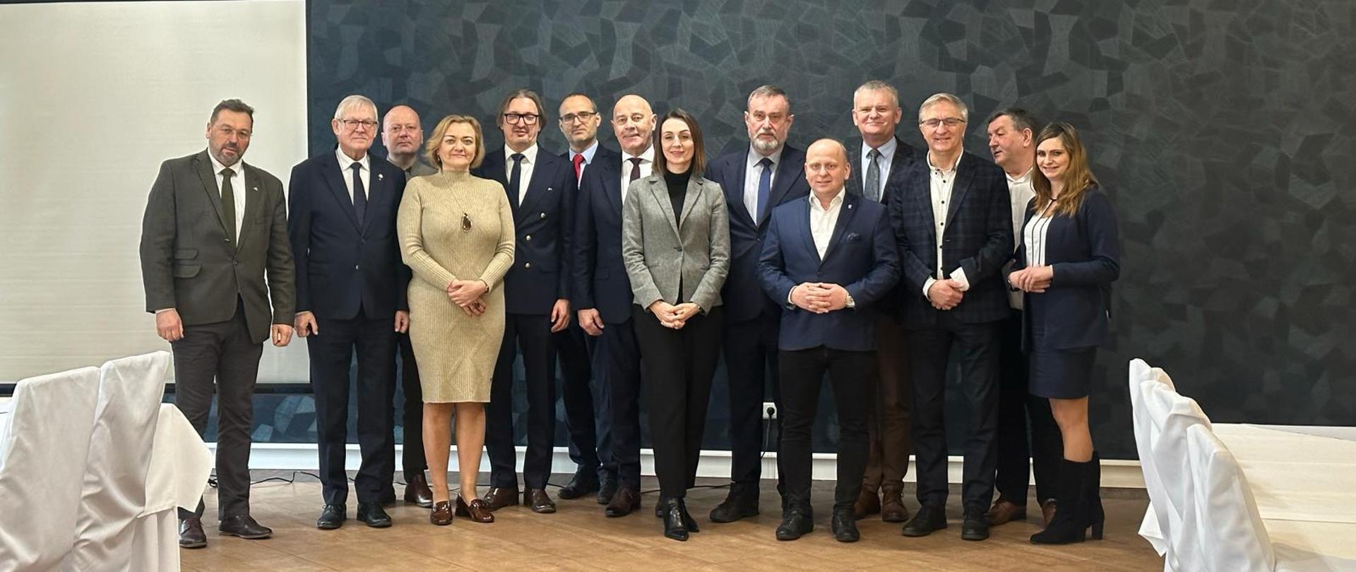 Grupa kilkunastu osób (samorządowców) pozuje do zdjęcia w dużej sali 