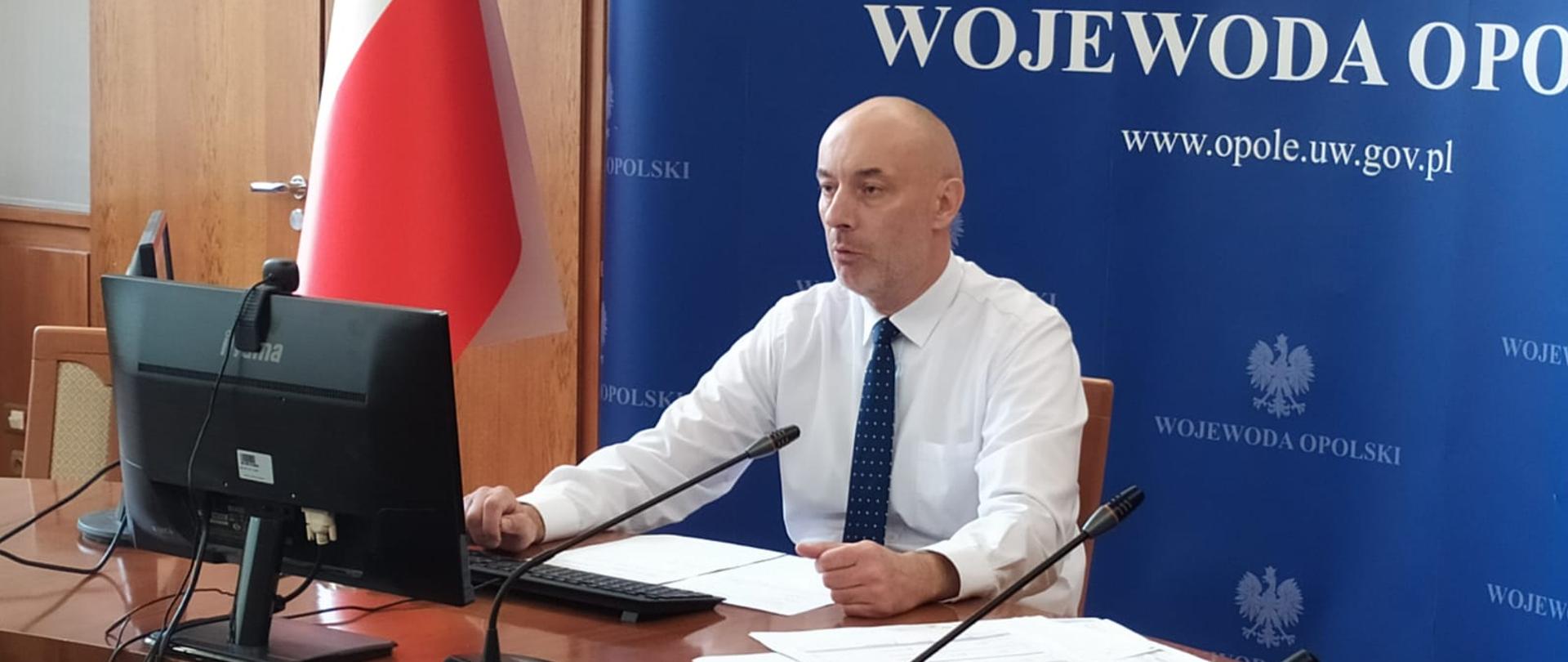 Wojewoda opolski podczas konferencji prasowej prowadzonej online, siedzi przed monitorami.