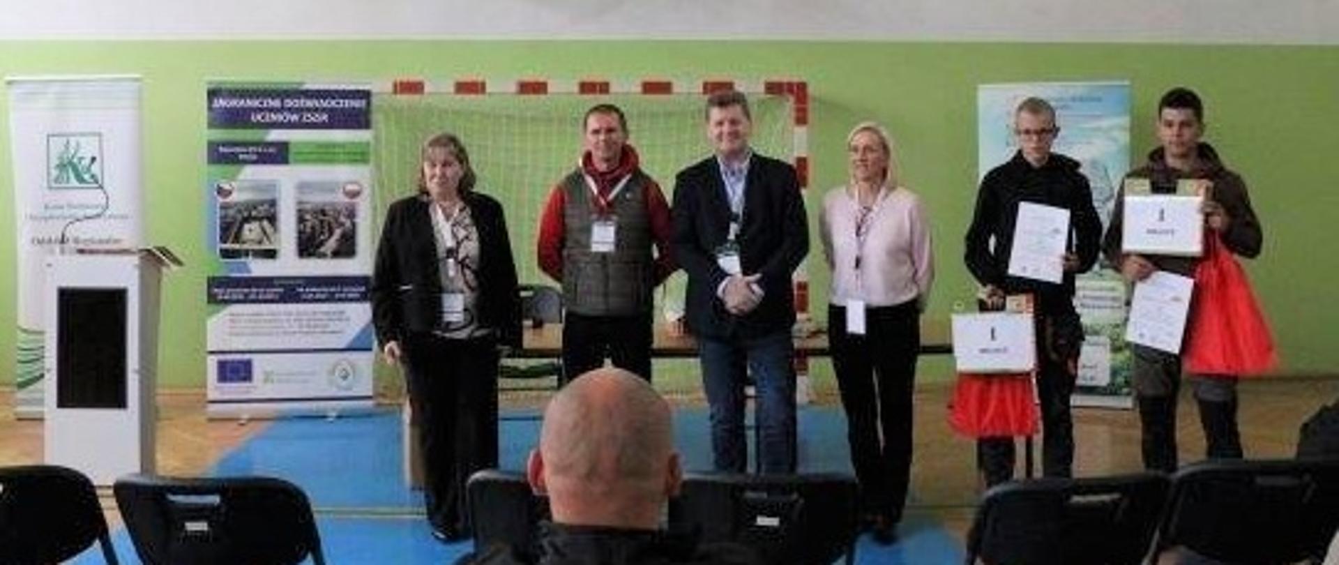 Na sali gimnastycznej stoją dwaj nastoletni chłopcy z dyplomami i nagrodami w rękach, po ich lewej stronie stoją dwie kobiety i dwaj mężczyźni z identyfikatorami na szyi. Za nimi znajdują roll-upy z logo instytucji. 