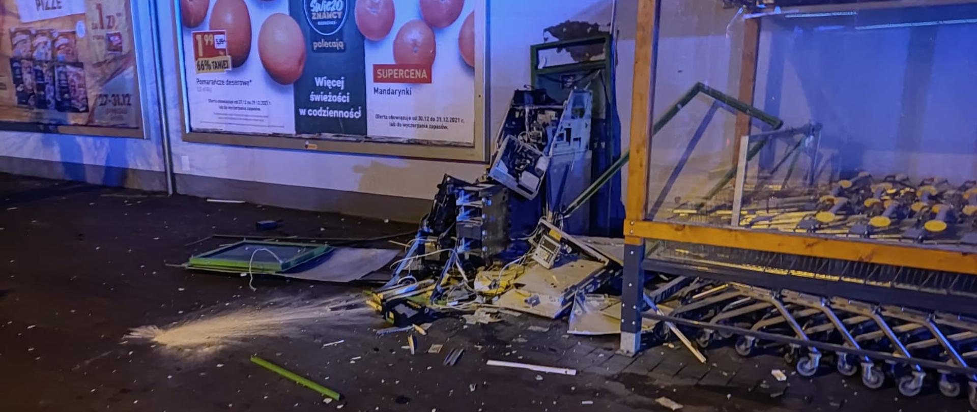 Fotografia przedstawia kompletnie zniszczony wybuchem bankomat znajdujący się przy markecie Biedronka, obok znajdują się wózki sklepowe. Na tylnej ścianie budynku widać banery reklamowe.